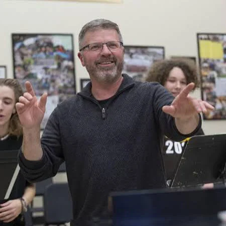Smiling conductor gestures with both hands while leading musicians during a concert band rehearsal.