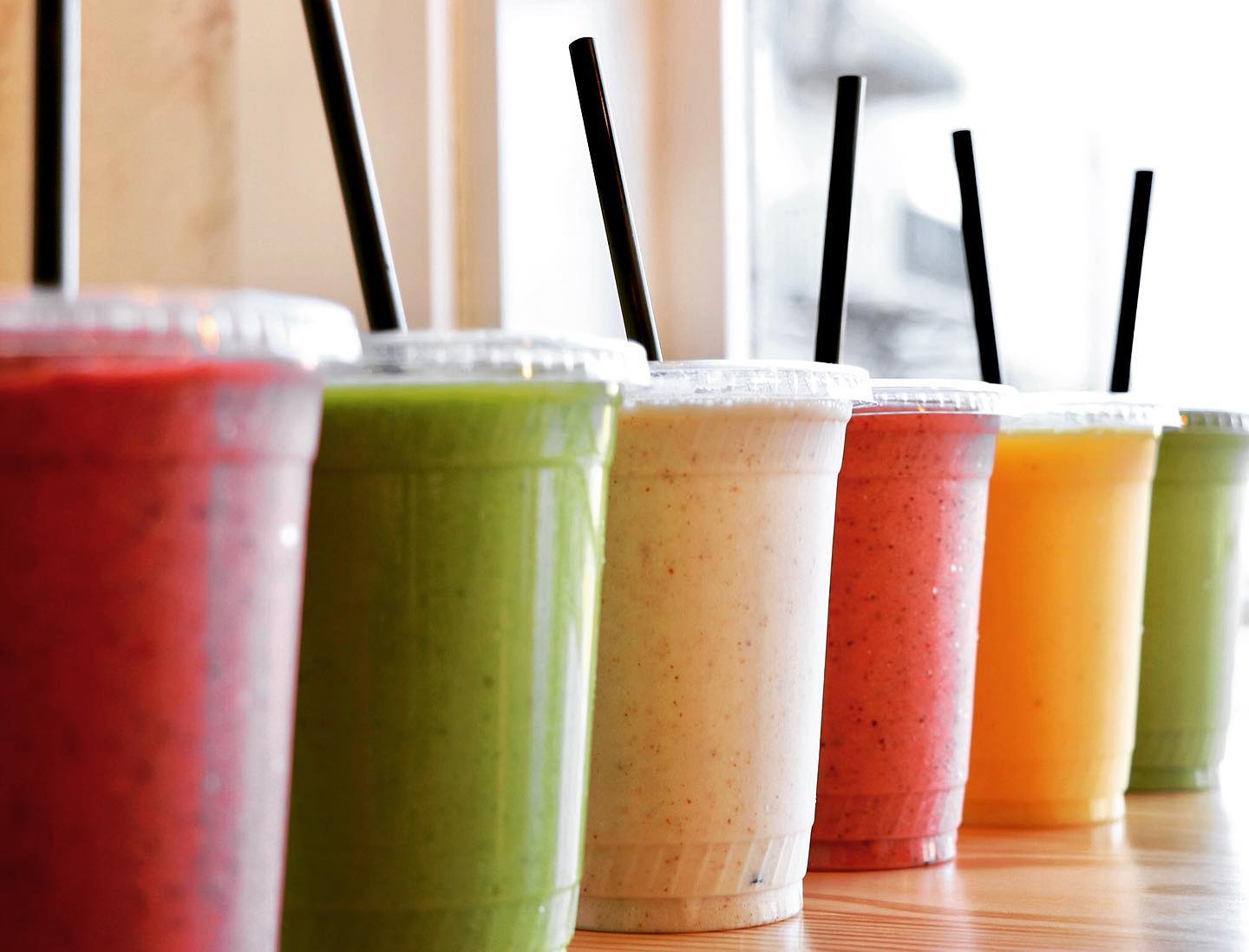 Foggy's Cafe colorful smoothies in clear cups with lids and black straws are lined up on a wooden counter, showcasing a variety of fresh fruit and vegetable blends.