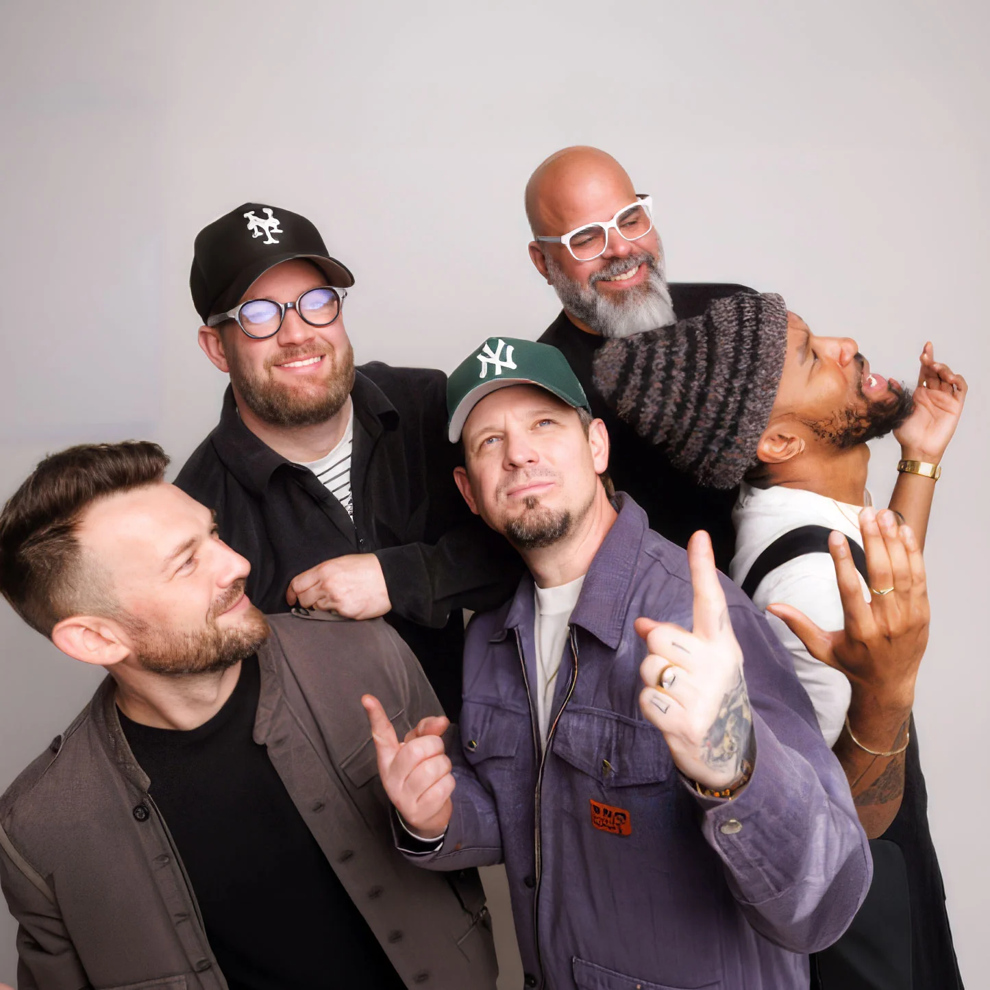 Five men pose playfully together against a light background, smiling and gesturing toward one another.