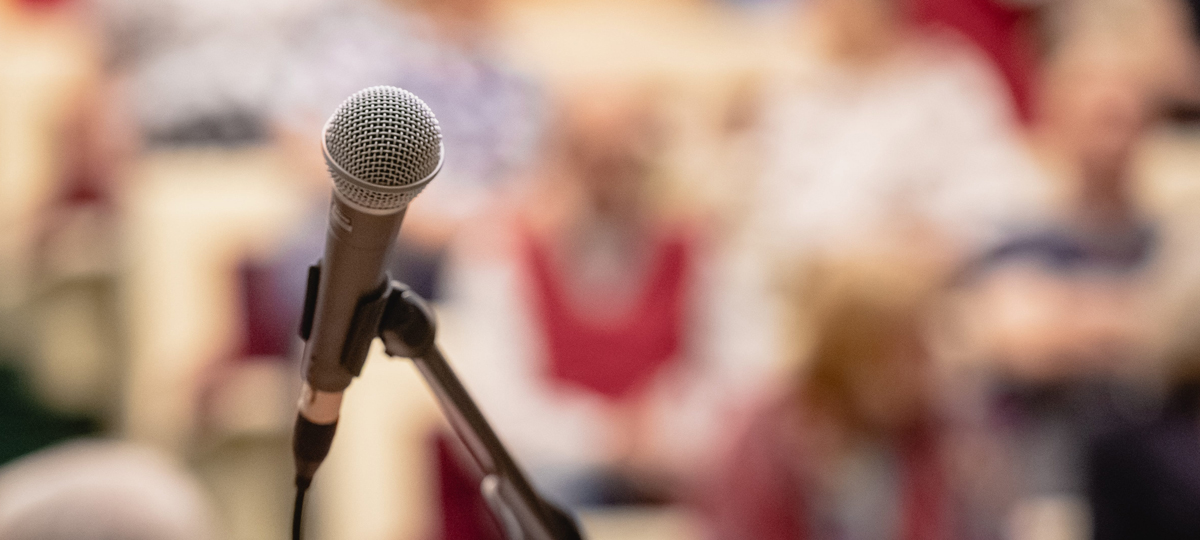 Microphone on a stand in sharp focus with a blurred audience seated in the background, suggesting a public talk, presentation, lecture, community meeting, or live event.