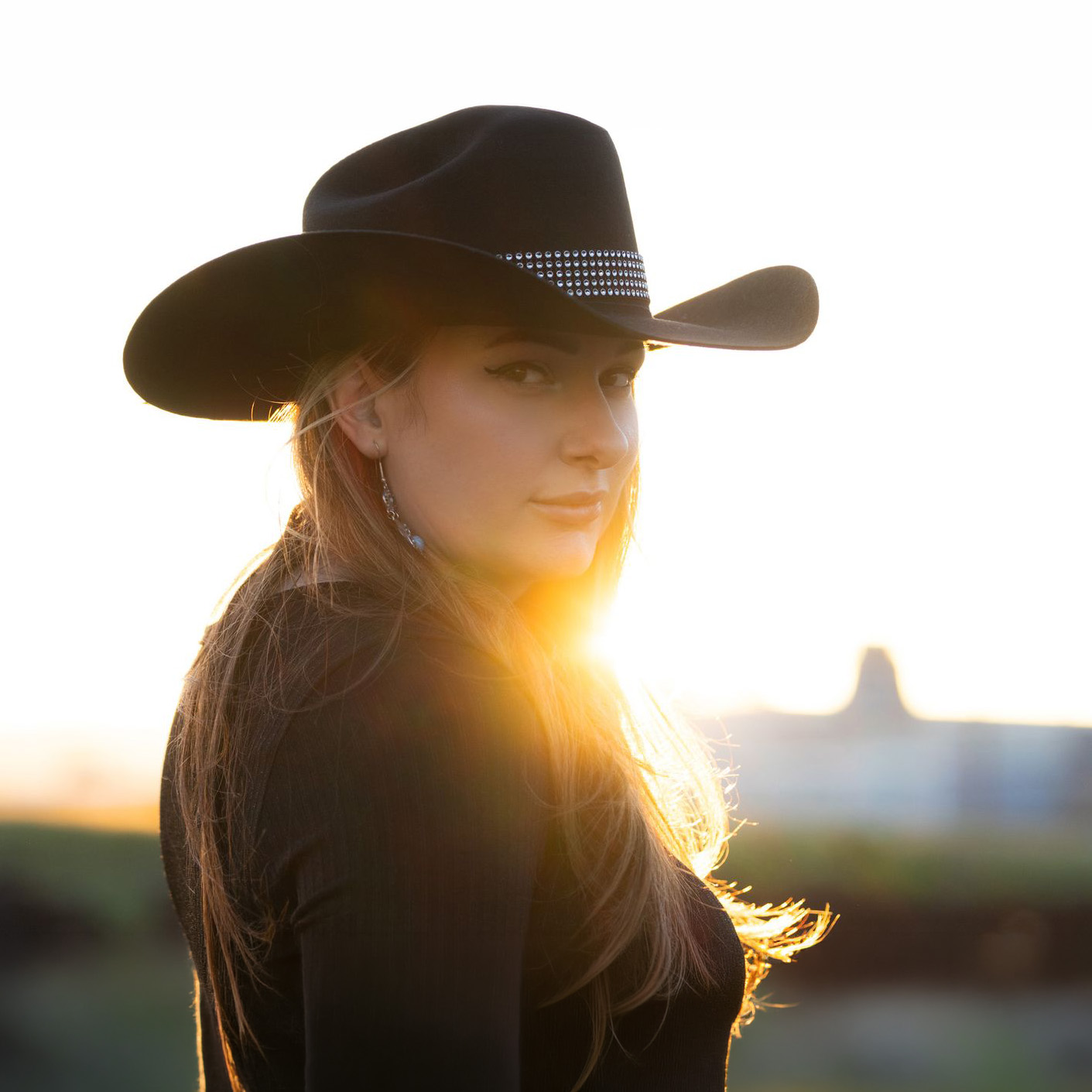 Person wearing a black cowboy hat and dark clothing looking over their shoulder toward the camera, backlit by a low sunset with a softly blurred outdoor landscape in the background.