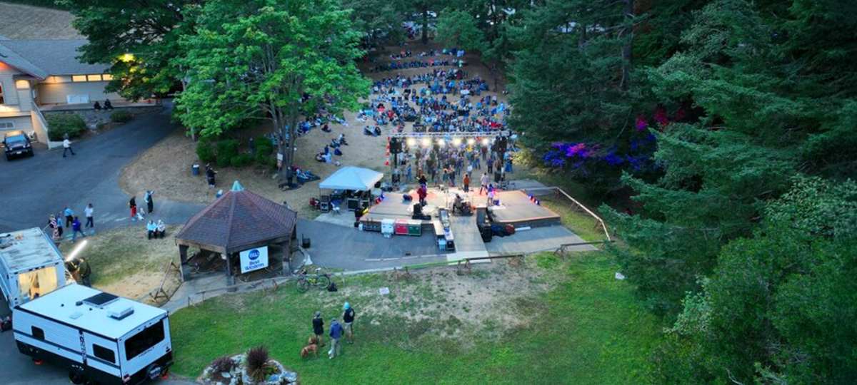 Aerial view of an outdoor concert venue surrounded by tall trees, with a crowd seated on a hillside facing a lit stage at dusk.