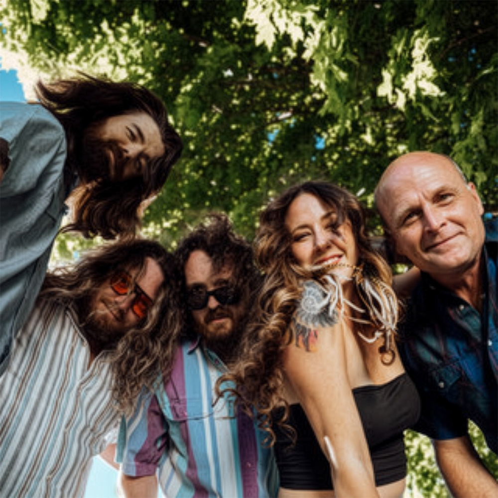 Five people leaning toward the camera outdoors beneath leafy trees, smiling and posing together in casual clothing, with sunlight filtering through the branches above.