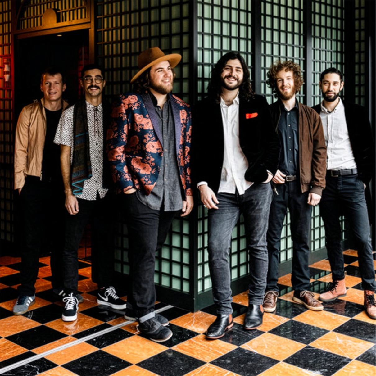 Six people standing together indoors on a black-and-gold checkered floor, wearing a mix of casual and dressy clothing, posing and smiling in front of decorative paneled walls.