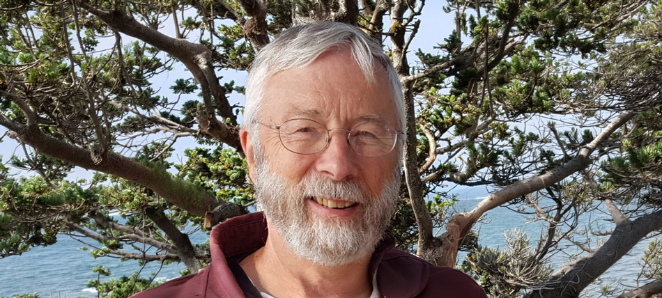 Portrait of a smiling man with a gray beard and glasses standing outdoors. Trees and coastal vegetation frame the background, with a glimpse of water visible beyond the branches.