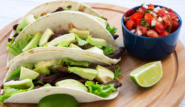 Los Guerreros Mexican Restaurant three soft tacos filled with grilled beef, avocado and lettuce, served with pico de gallo and fresh lime wedges on a wooden board.