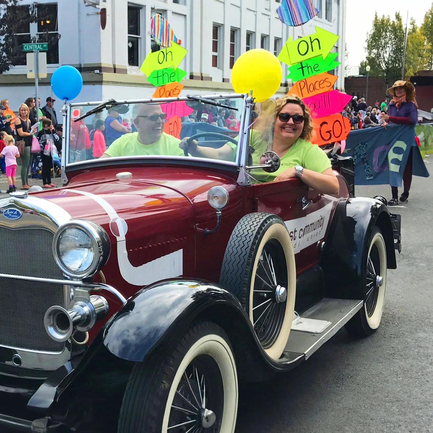 Vintage red convertible in a community parade with two smiling riders wearing neon green shirts. Bright balloons and colorful "Oh, the Places You'll Go!" signs decorate the car as crowds watch.