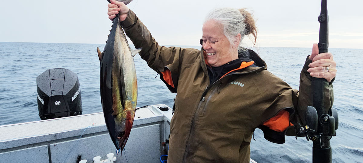 Woman on a fishing boat in open ocean waters, smiling while holding a freshly caught tuna in one hand and a fishing rod in the other. Overcast sky and calm sea in the background.
