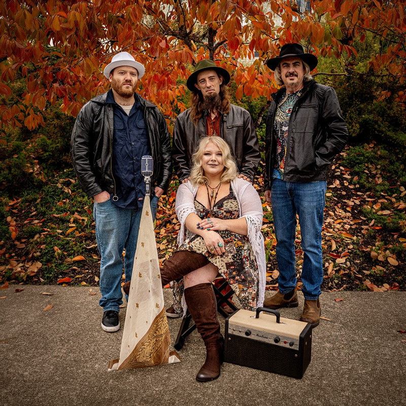 Four musicians posing outdoors in front of autumn foliage, with one seated and three standing beside musical equipment including a microphone and amplifier.