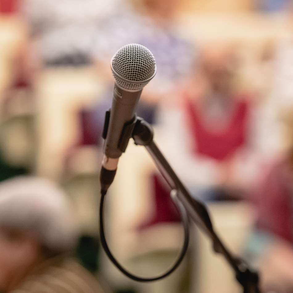 Microphone on a stand in sharp focus with a blurred audience seated in the background, suggesting a public talk, presentation, lecture, community meeting, or live event.