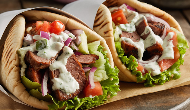 two pitas fill wrapped in paper, filled with shaved beef, veggies and tzatziki sauce