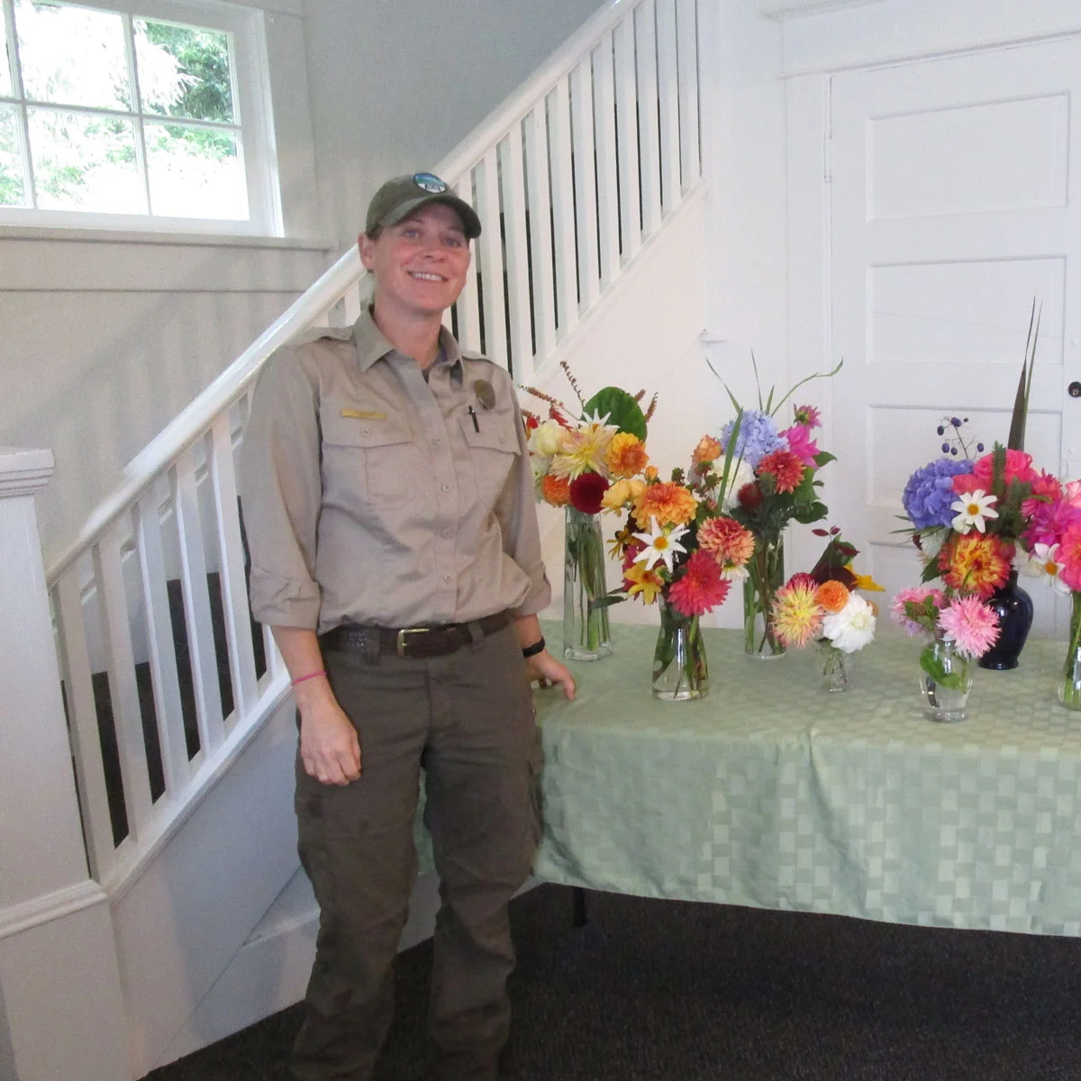 Park ranger standing beside a table displaying colorful flower arrangements inside a historic building with a staircase and large window in the background.