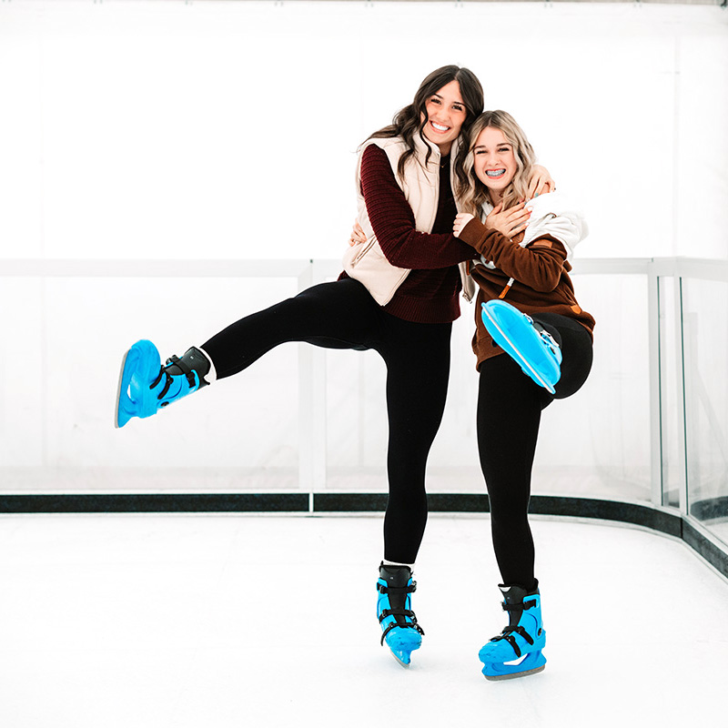 two young women wearing leggings, cosy tops and bright blue ice skates on an ice rink