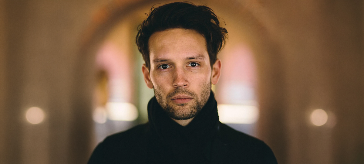 A close-up portrait of a person with dark, styled hair and light facial stubble, wearing a black high-collared coat, looking directly at the camera with a neutral expression against a blurred background.