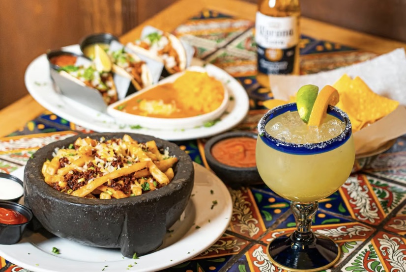 variety of Mexican dishes atop a table next to a Margarita and Corona beer
