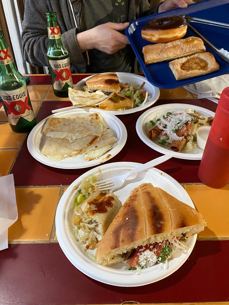 four plates of Mexican food on a table next to two bottle of Dos Equis beer and a tray of doughnuts