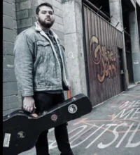 Gabriel John holding his guitar case on the street
