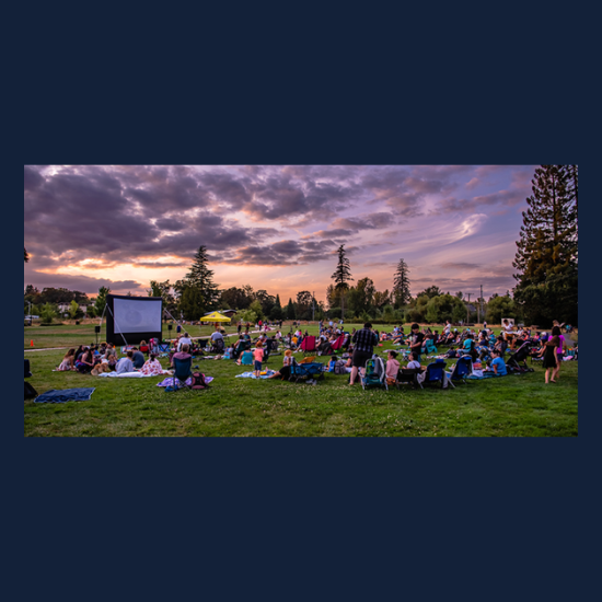 A large, inflatable film screen sits in a sunset-lit park with a crowd of people in front of it.