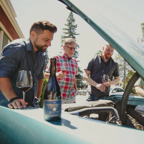 People looking into a car hood with wine in hand