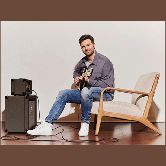 Jackson Michaelson in a chair playing a guitar that is also plugged into an amp