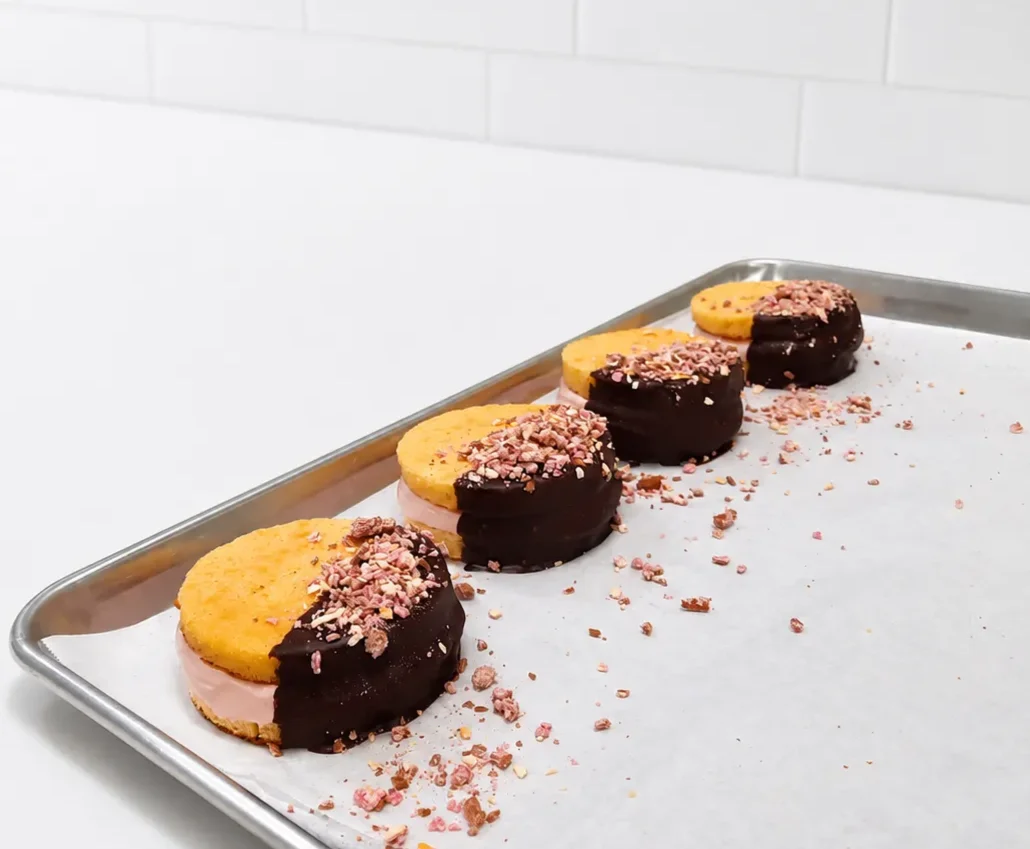 A baking tray holding four ice cream sandwiches, each partially coated in dark chocolate and pink sprinkles.