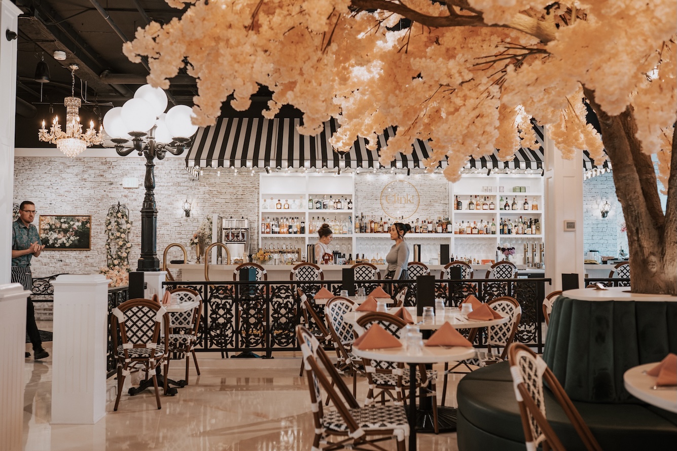 restaurant interior designed as a French cafe with woven chairs, round white tables, white brick wall, lamp post and tree with pink foliage