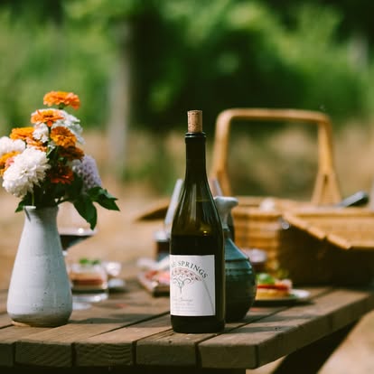 Outdoor scene of a table with picnic basket, white vase of orange and white flowers, foods, and a bottle of Vitae Springs wine