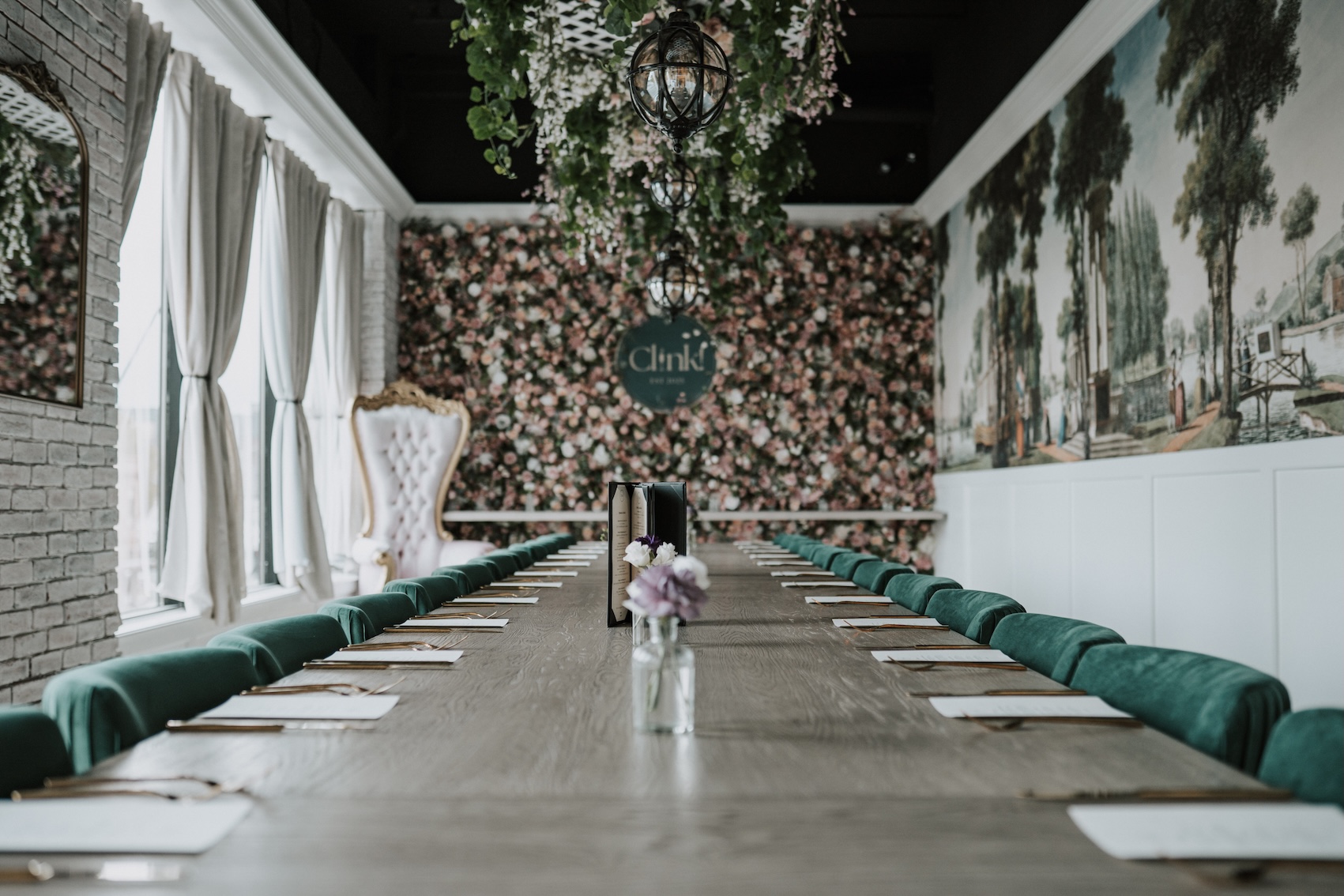 Private dining space with wooden table, green cloth chairs, wall of windows, wall of flowers, and wall with mural of cityscape.