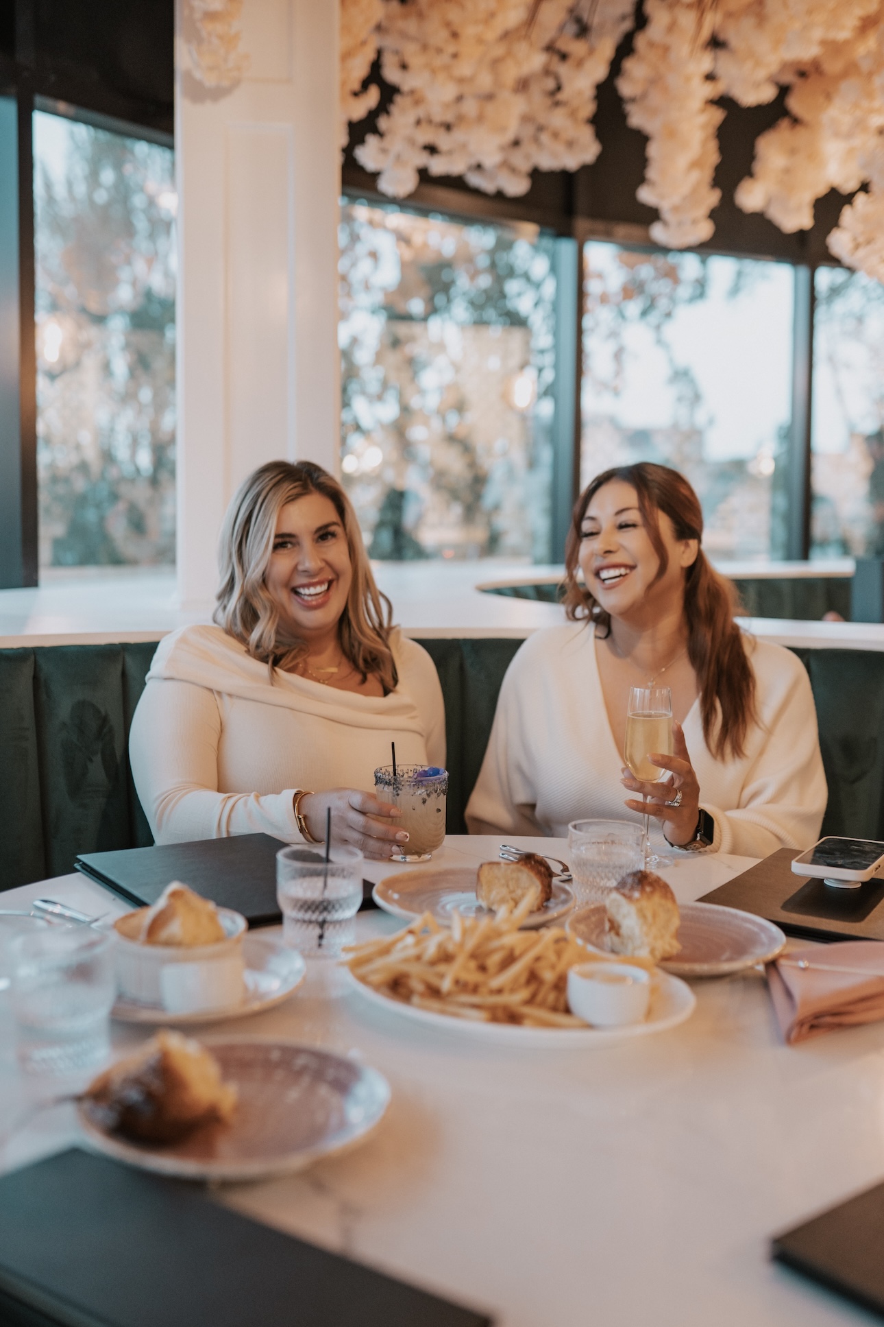 two women wearing white sit at a table with food and drinks