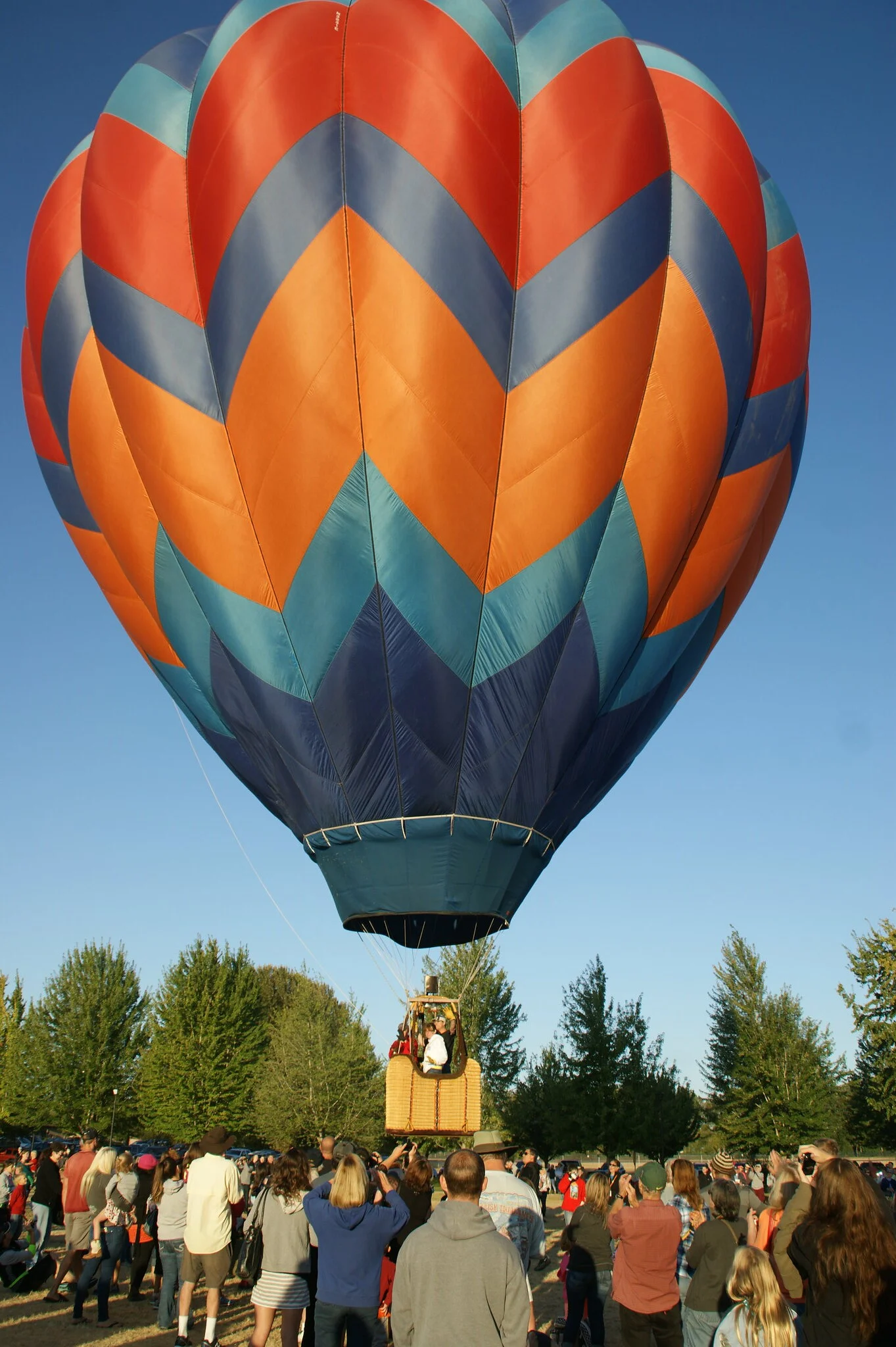 Crowd of people watch a blue and orange chevron striped hot air balloon taking off on a sunny day