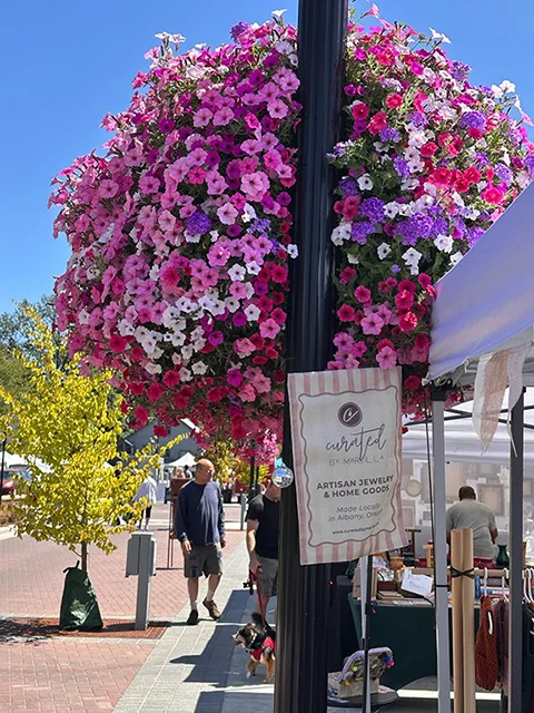 A couple of adult men browsing booth at the artisan faire with a small dog on a sunny day. A large flower hanging basket of pink, purple, and white flowers in the foreground