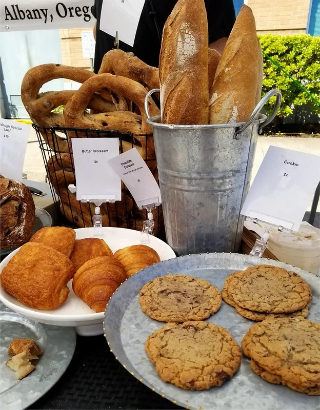 Market booth display of home baked goods including cookies, butter croissants, soft pretzels, and baguettes