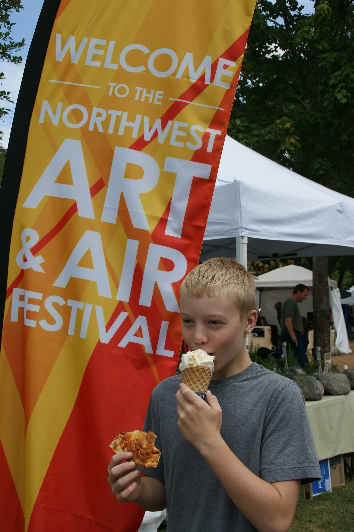 Young boy in grey shirt eating a vanilla ice cream cone and pizza in front of Northwest Art & Air Festival banner