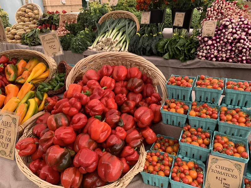 Market booth full of fresh and brightly colored produce including cherry tomatoes, red bell peppers, radishes, green onions, and various hot pepper varieties