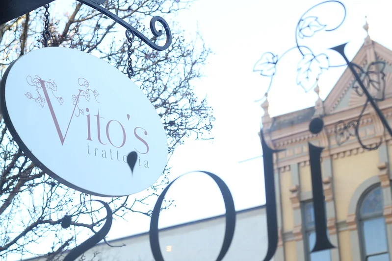 Window shot of Vito's Trattoria's hanging sign displaying their logo in a sophisticated red font.