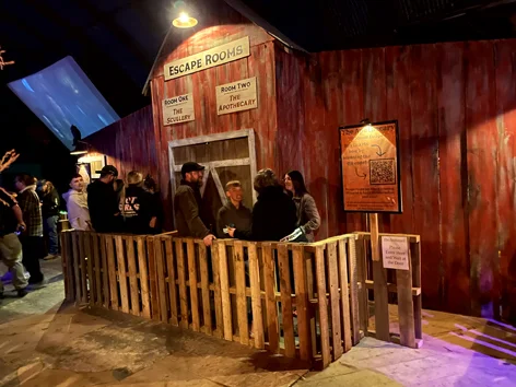 A line of people wait ouside a old leaning red bard to participate in an escape room.