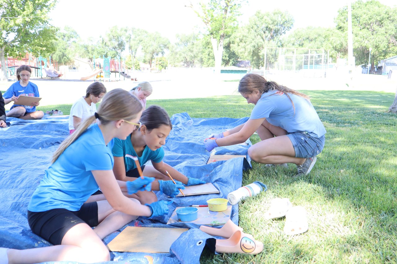 Kids sit on a blue tarp in a grass field while painting miniature murals.