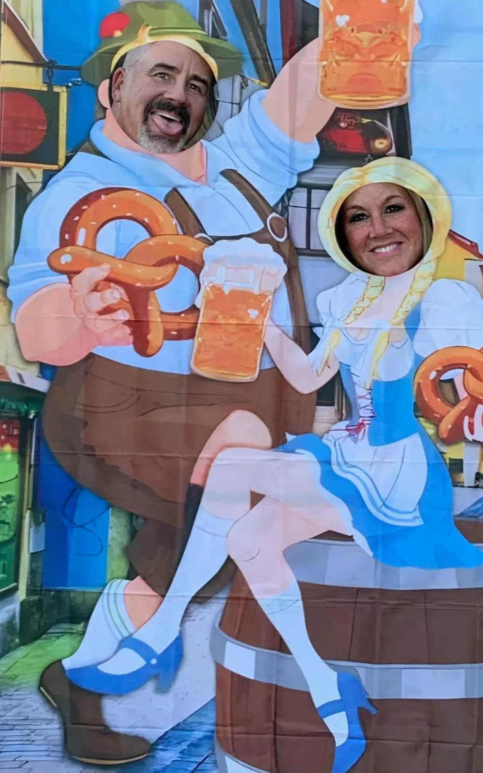 Two people pose behind a cutout that shows European figures eating pretzels and drinking beer.