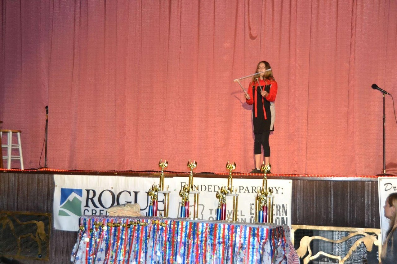 A girl juggles sticks on stage while an array of trophies stand on a table in the foreground.